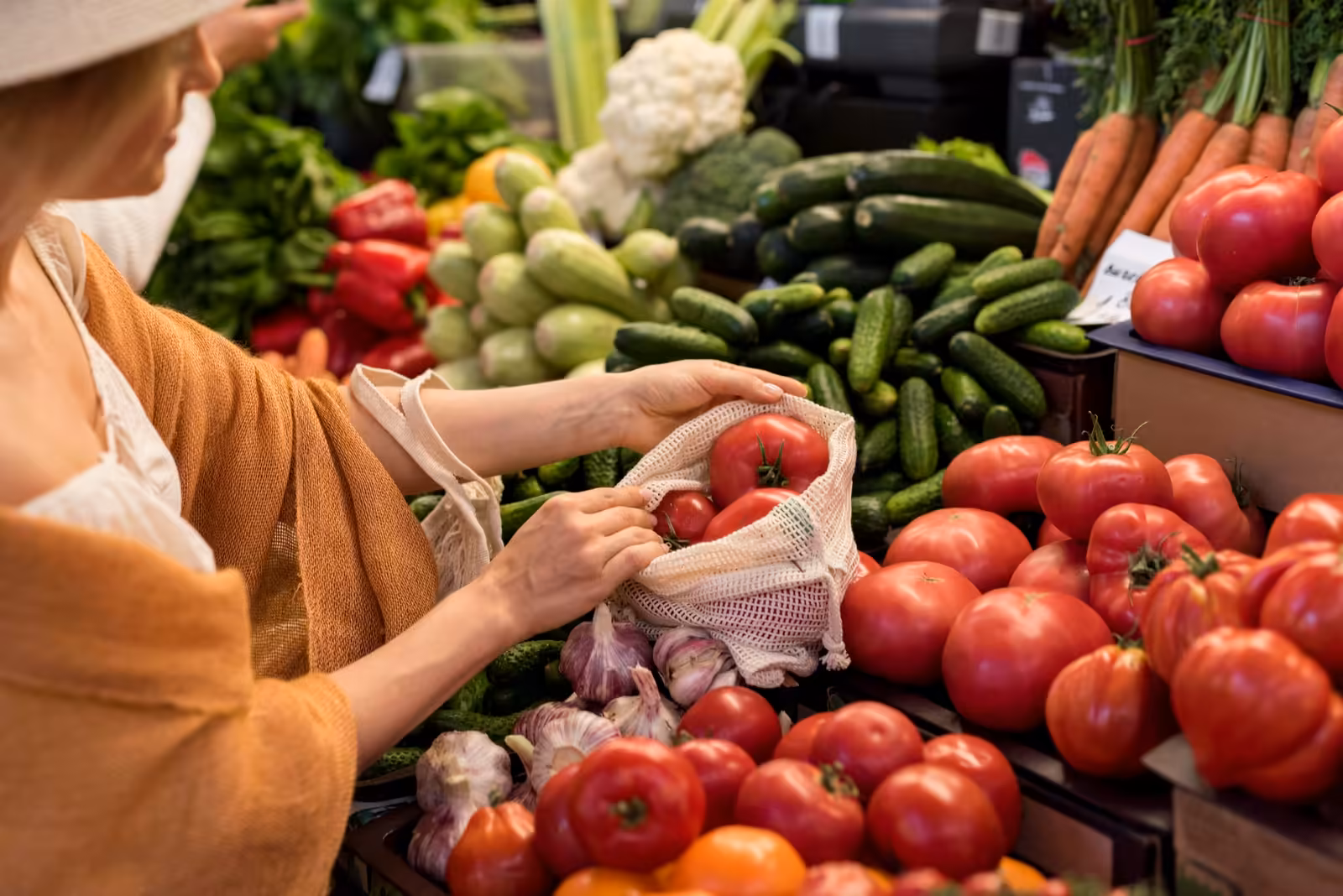 Étal de marché avec tomates, concombres et ail, une cliente remplit son sac en filet